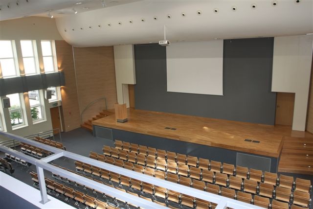 Sala konferencyjna z drewnianą podłogą i rzędami krzeseł, widok z góry z balkonu, z widoczną sceną i projektorem.