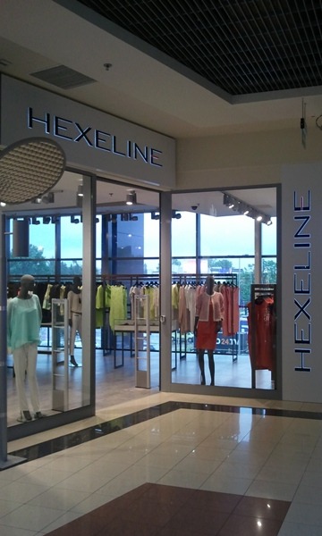 Wejście do sklepu odzieżowego Hexeline z wystawą manekinów w ubraniach w pastelowych kolorach i błyszczącą podłogą z płytek.