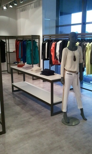 Jasnoszara posadzka imitująca beton w nowoczesnym sklepie odzieżowym, z ekspozycją ubrań na wieszakach i manekinie w białym swetrze i spodniach.