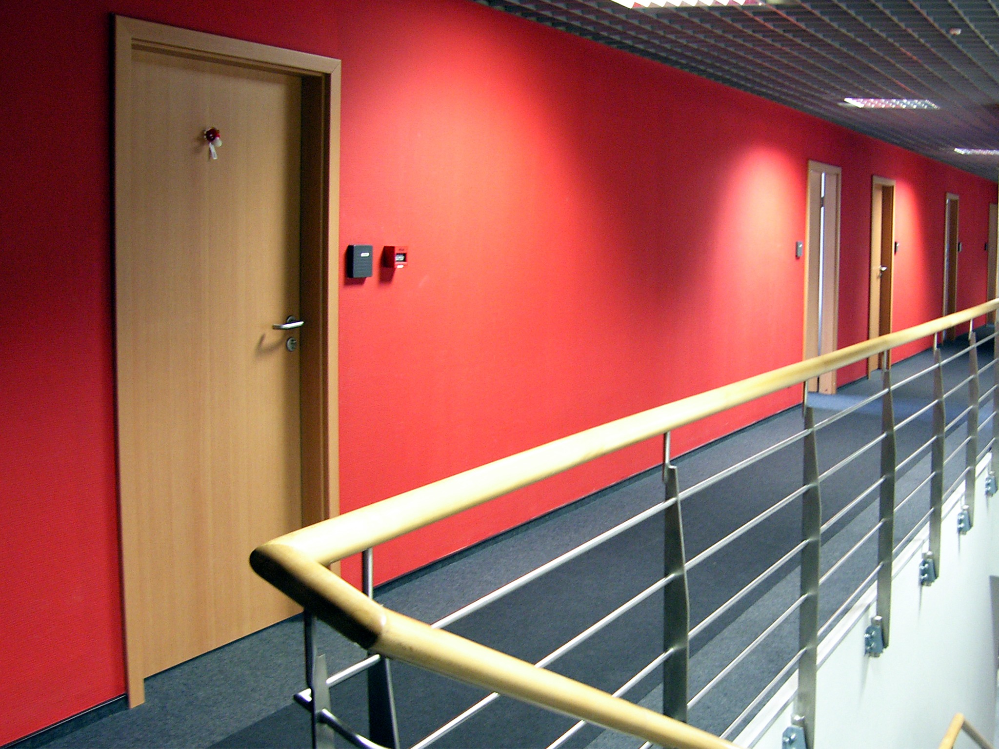 Korytarz biurowy z czerwonymi ścianami, szarą wykładziną dywanową i drewnianymi drzwiami, widok z góry schodów z balustradą.