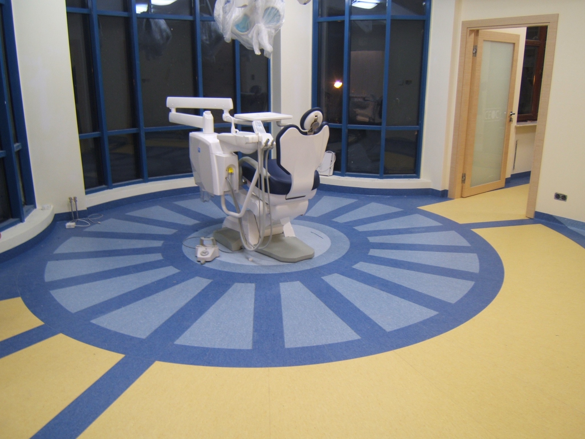 Wnętrze gabinetu lekarskiego z krzesłem dentystycznym, podłoga z wykładziny w geometryczny wzór z przewagą koloru niebieskiego i żółtego, duże okna z ciemnymi szybami.