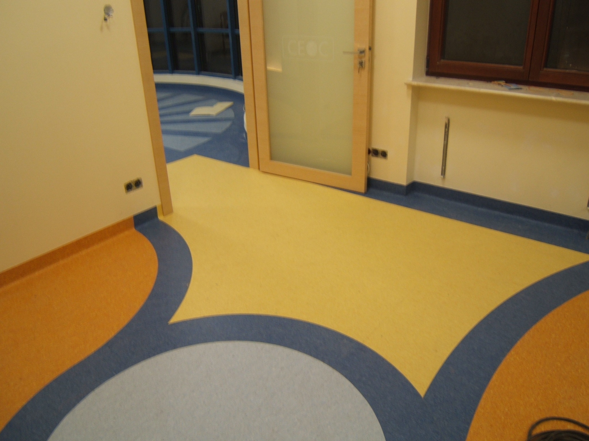 Wielokolorowa podłoga z wykładziny PCV w kształcie fal, z widocznymi detalami łączeń i wzorów, w pomieszczeniu z oknem i drzwiami z matową szybą.