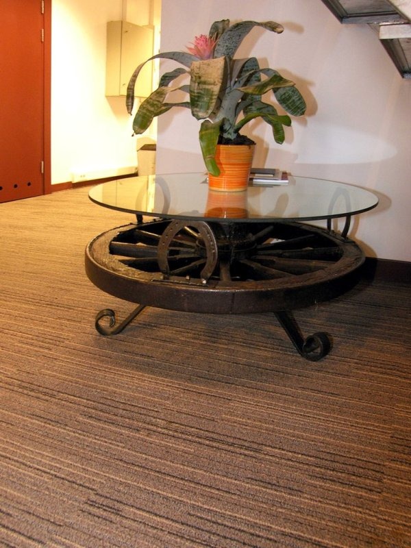 Brązowa wykładzina dywanowa z subtelnym wzorem w paski, widoczny fragment szklanego stolika kawowego z podstawą z koła od wozu i rośliną doniczkową na blacie.