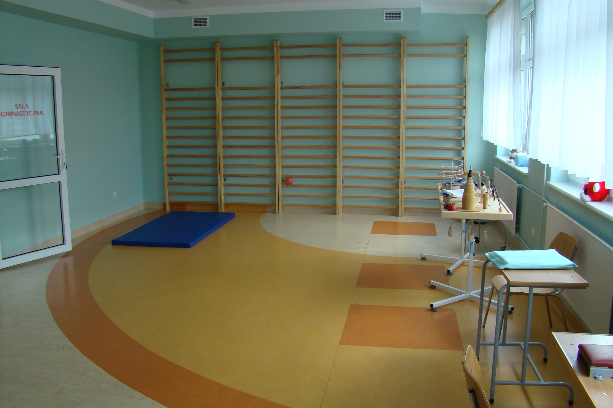 Sala gimnastyczna z drewnianymi drabinkami, podłogą w odcieniach beżu i brązu, niebieskim materacem i stolikami z przyborami do ćwiczeń.