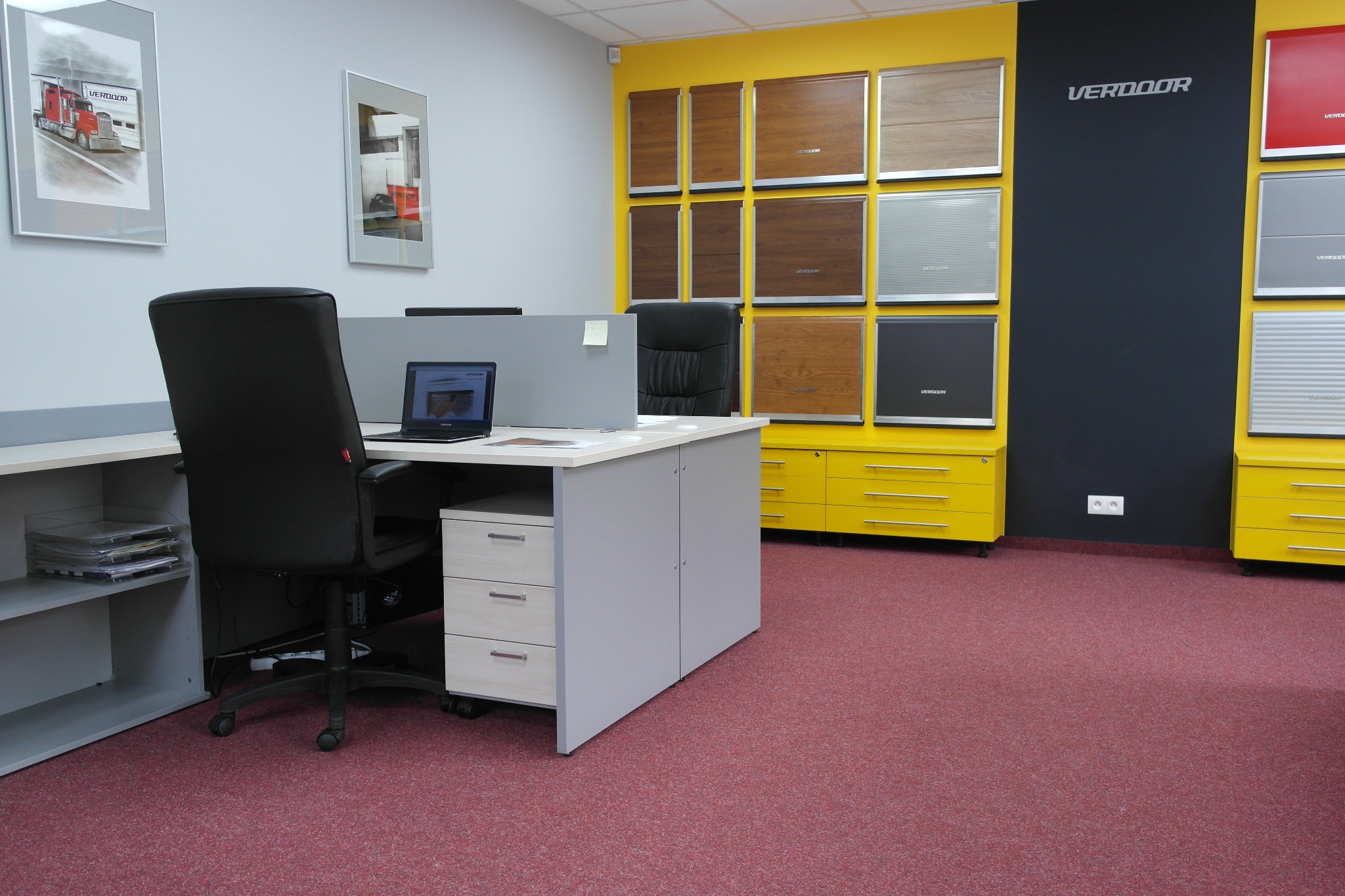 Biuro z ekspozycją próbek różnych typów bram garażowych: drewnianych, metalowych, roletowych; na podłodze czerwona wykładzina dywanowa.