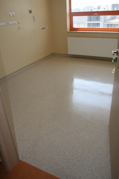 Jasna, pusta przestrzeń z nową, jednolitą powierzchnią podłogową z drobnym wzorem, widok na okno z pomarańczową ramą i grzejnik.