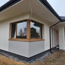 Elevaterm - Nowoczesny dom jednorodzinny z jasną elewacją, drewnianymi oknami i szarą okładziną cokołu, widoczne przewody wystające ze ściany, pracownik siedzący na zewnątrz budynku.