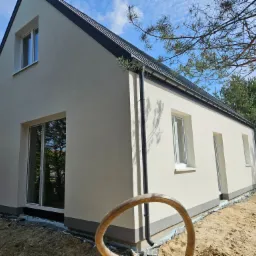 Świeżo otynkowany dom jednorodzinny z czarną dachówką, widok z zewnątrz, w tle drzewa. Wokół domu piasek i fragment węża ogrodowego.