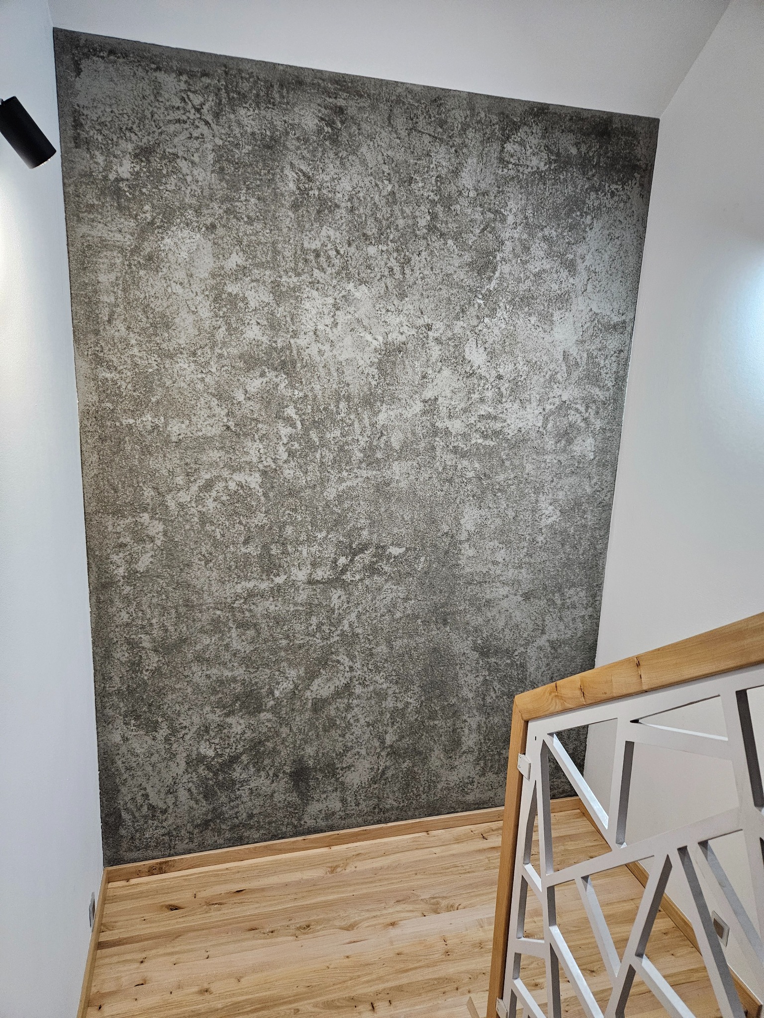 Dekoracyjna ściana o fakturze betonu w klatce schodowej, widoczny drewniany parkiet i fragment białej, ażurowej balustrady.