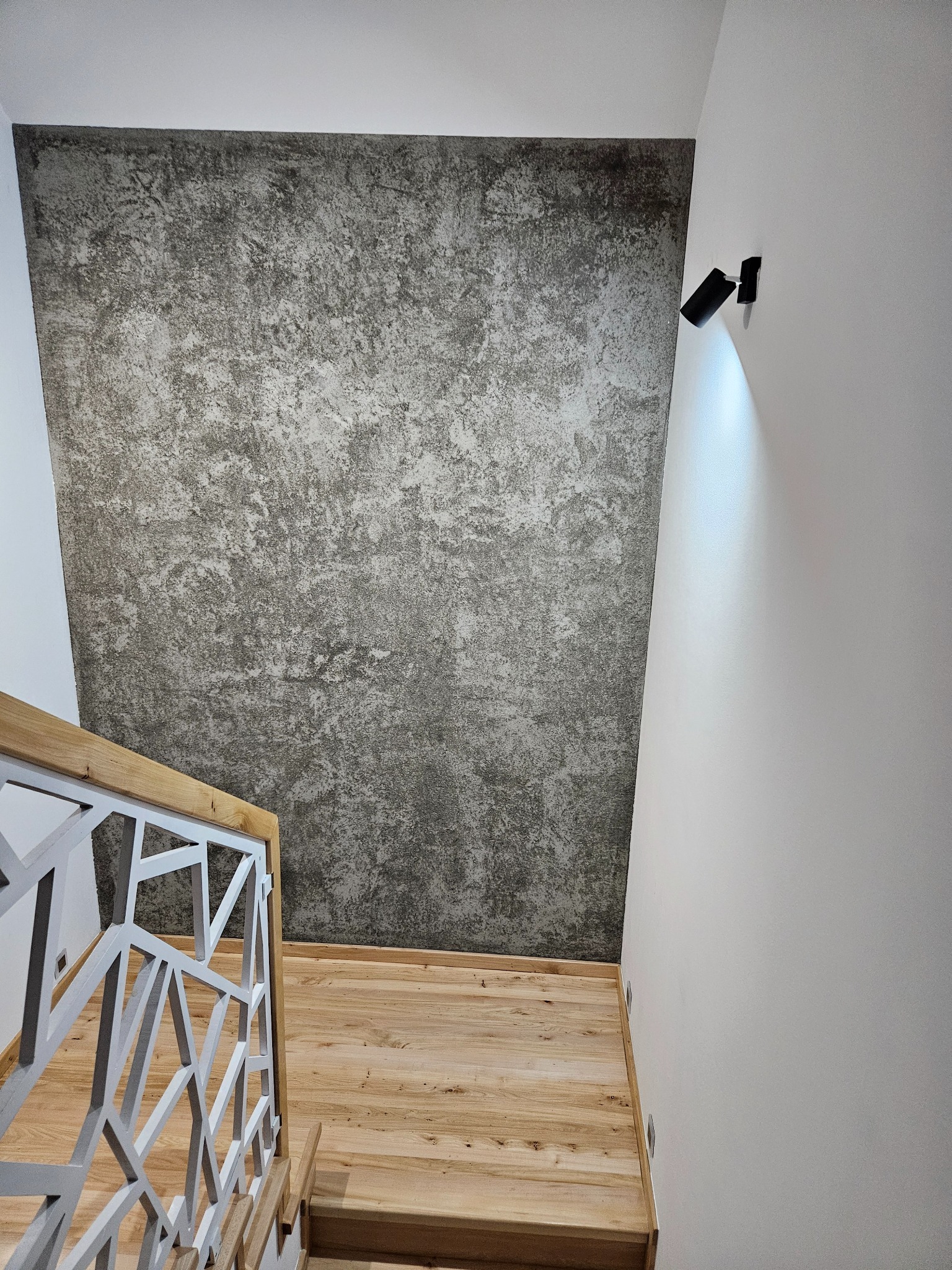 Ściana z efektem betonu architektonicznego, nowoczesne oświetlenie punktowe i drewniane schody z białą, geometryczną balustradą.
