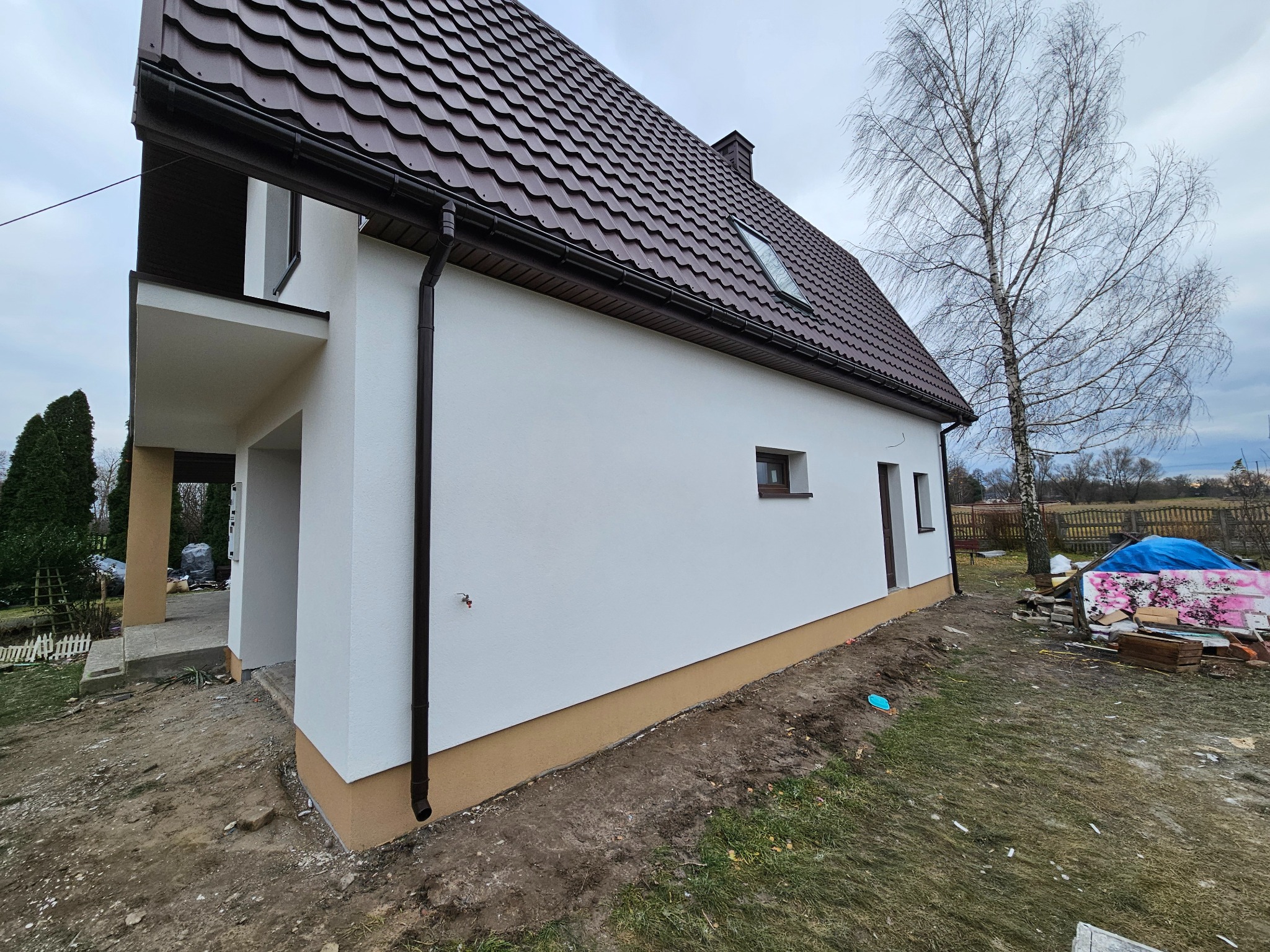 Świeżo otynkowana elewacja domu jednorodzinnego z brązowym dachem i rynnami, widok z boku na tle nagiego drzewa i placu budowy z materiałami pod plandeką.