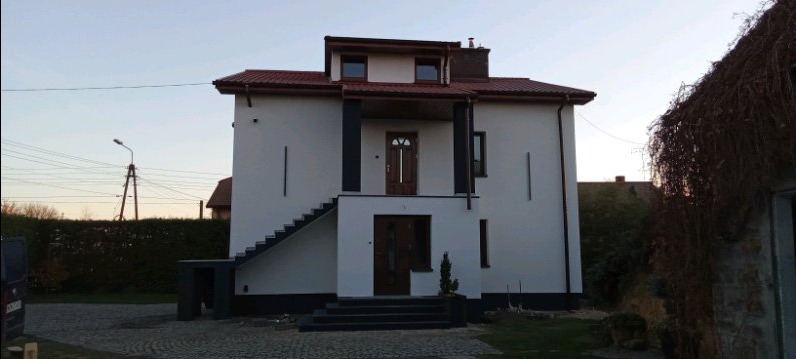 Biały, dwupiętrowy dom z ciemną stolarką okienną i drzwiową oraz schodami zewnętrznymi, widoczny wczesnym wieczorem.