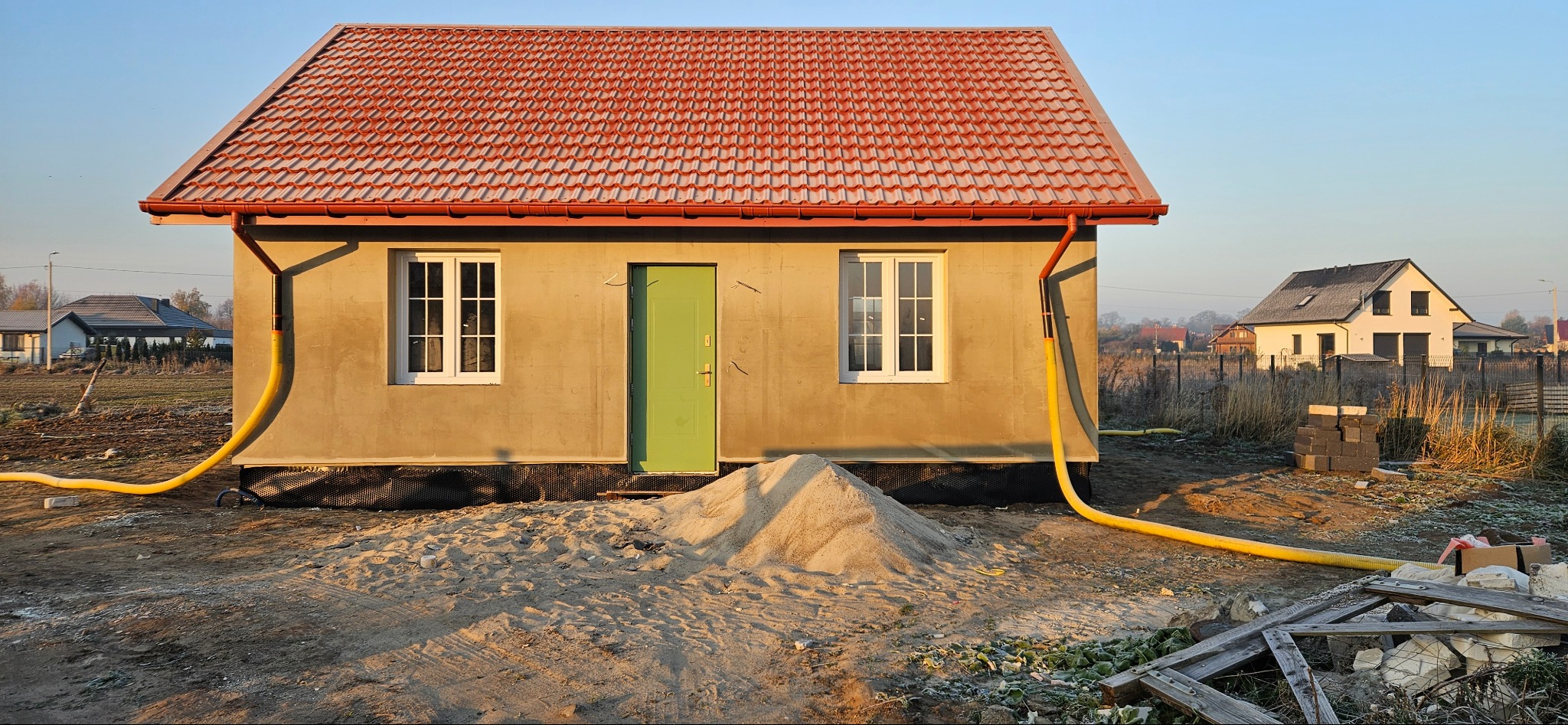 Dom w stanie surowym w Baranowie z czerwonym dachem i zielonymi drzwiami, widoczny stos piasku i żółte rury na tle porannego światła.