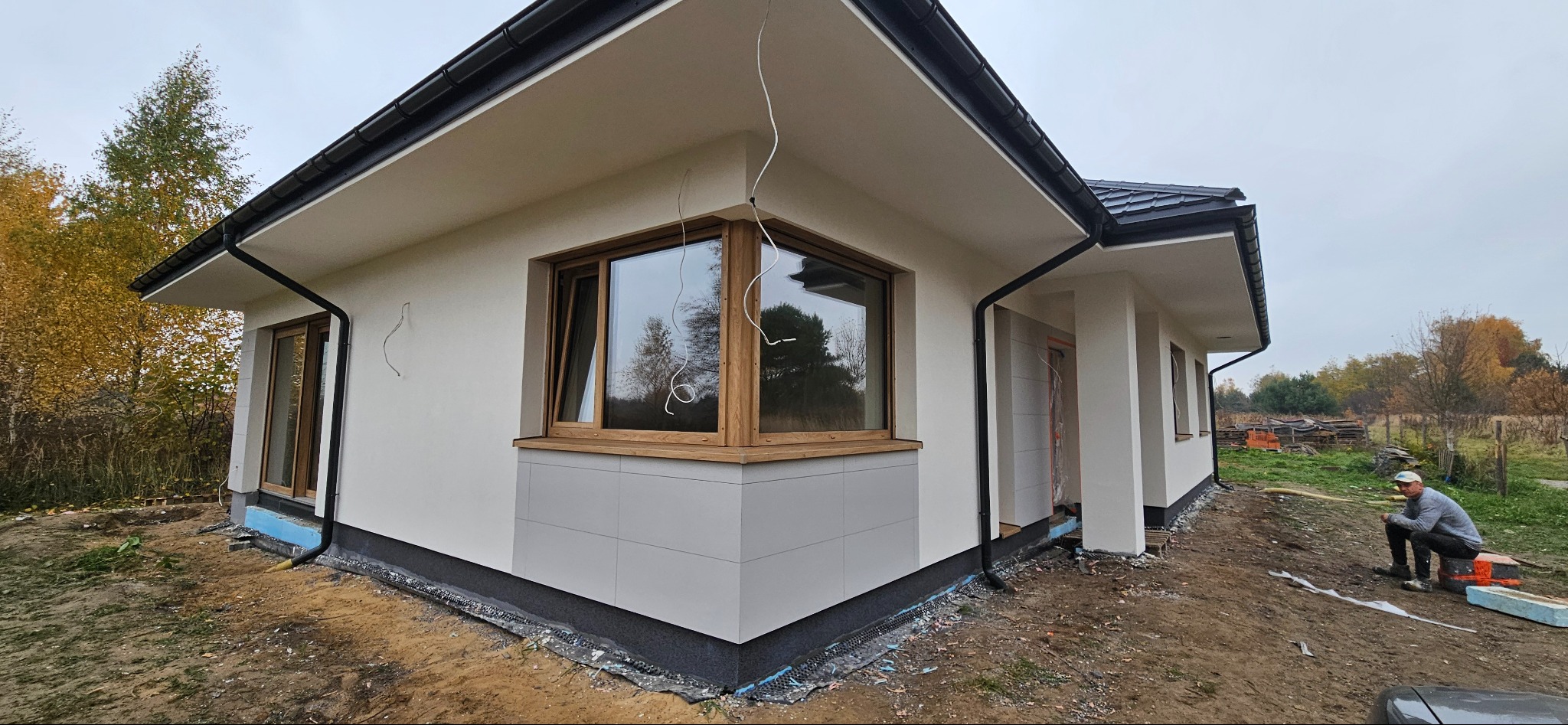 Nowoczesny dom jednorodzinny z jasną elewacją, drewnianymi oknami i szarą okładziną cokołu, widoczne przewody wystające ze ściany, pracownik siedzący na zewnątrz budynku.