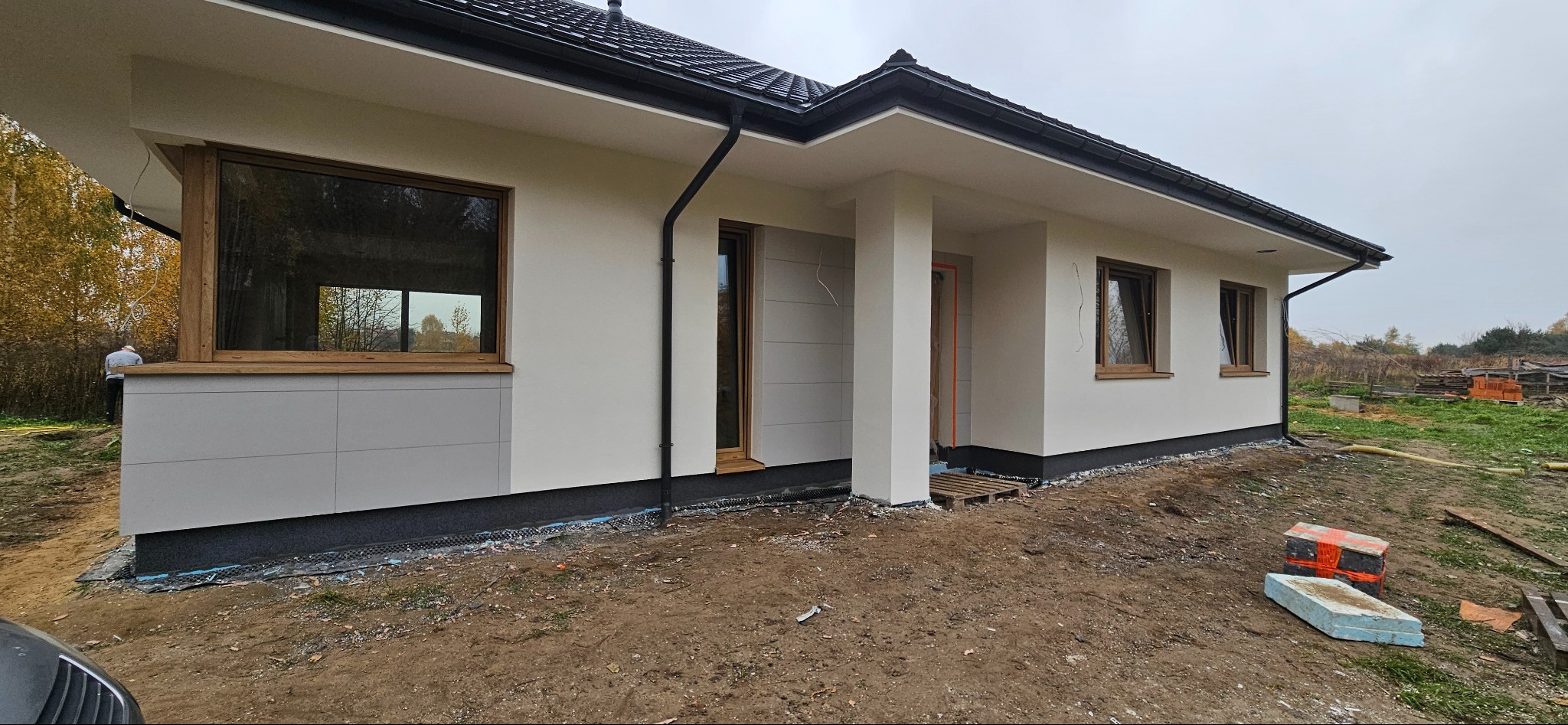 Nowoczesny dom jednorodzinny z jasną elewacją i drewnianymi oknami, widoczny z boku, z elementami wykończeniowymi w kolorze szarym, teren wokół domu w trakcie prac ziemnych.