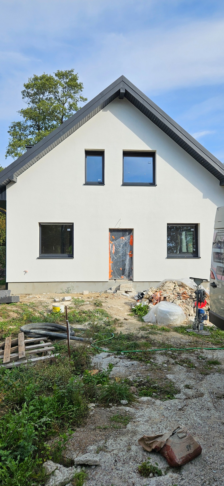 Nowo otynkowana elewacja domu jednorodzinnego z zamontowanymi oknami, zabezpieczone drzwi wejściowe folią, widoczne materiały budowlane i sprzęt na zewnątrz.