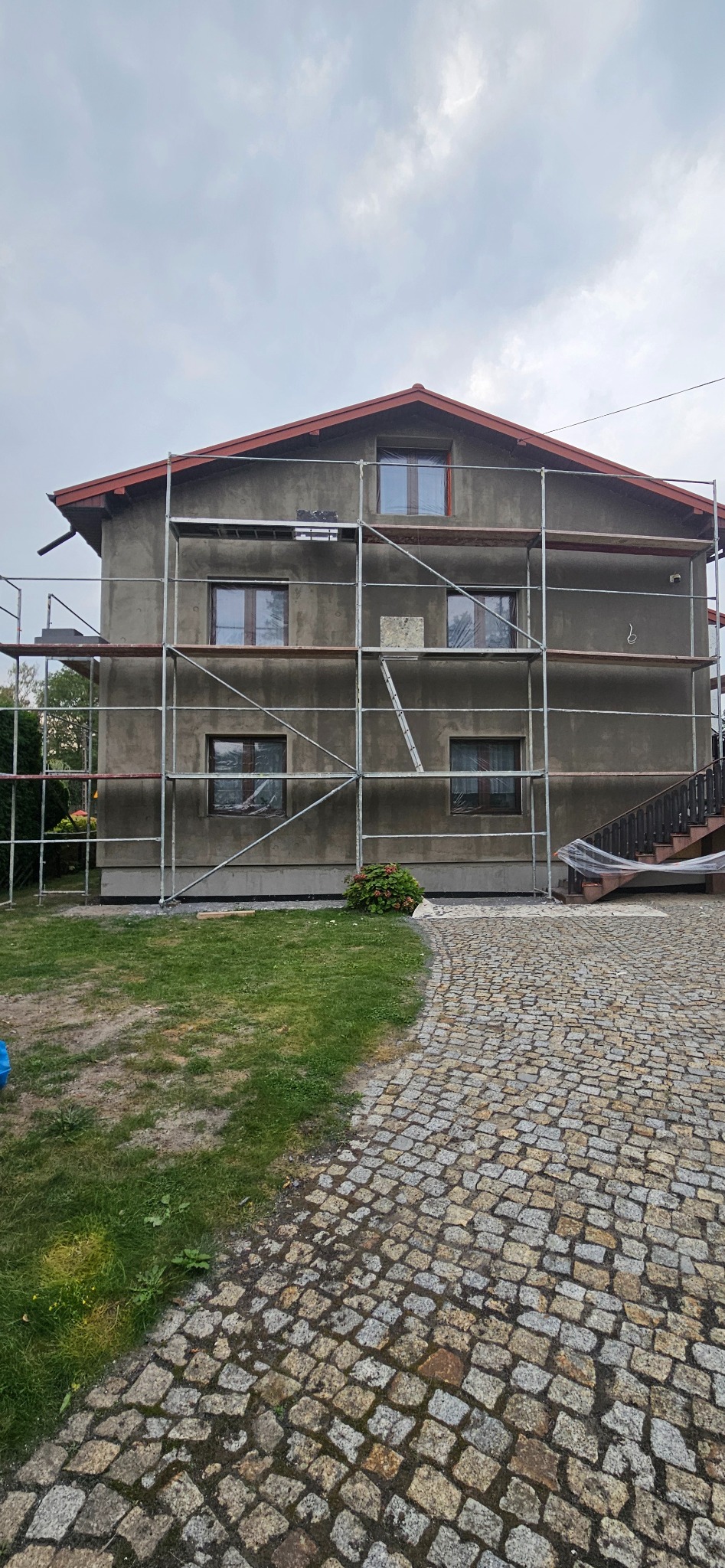 Dom jednorodzinny w trakcie ocieplania, z widocznym rusztowaniem i oknami zabezpieczonymi folią, na pierwszym planie trawnik i bruk z kostki.