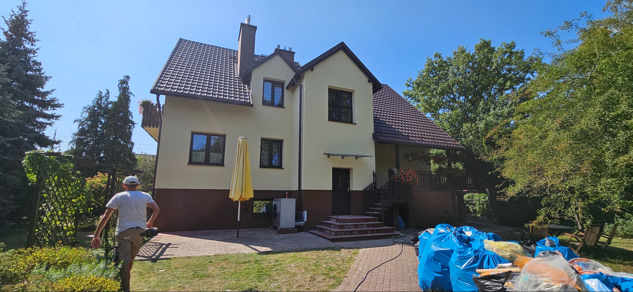 Dwukondygnacyjny dom z elewacją w odcieniach beżu i brązu, brązowy dach, widoczne prace porządkowe po remoncie, worki na śmieci, mężczyzna w oddali.