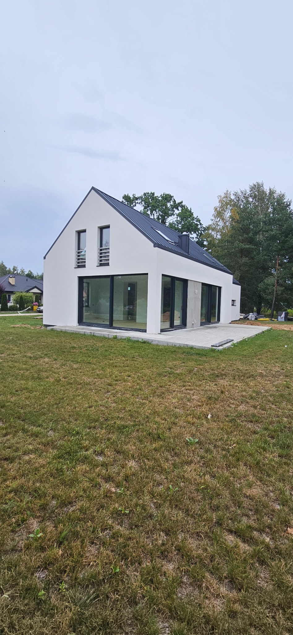 Nowoczesny dom jednorodzinny z białymi ścianami, czarnym dachem i dużymi oknami tarasowymi, widok z zewnątrz na trawnik i częściowo wykończony taras.