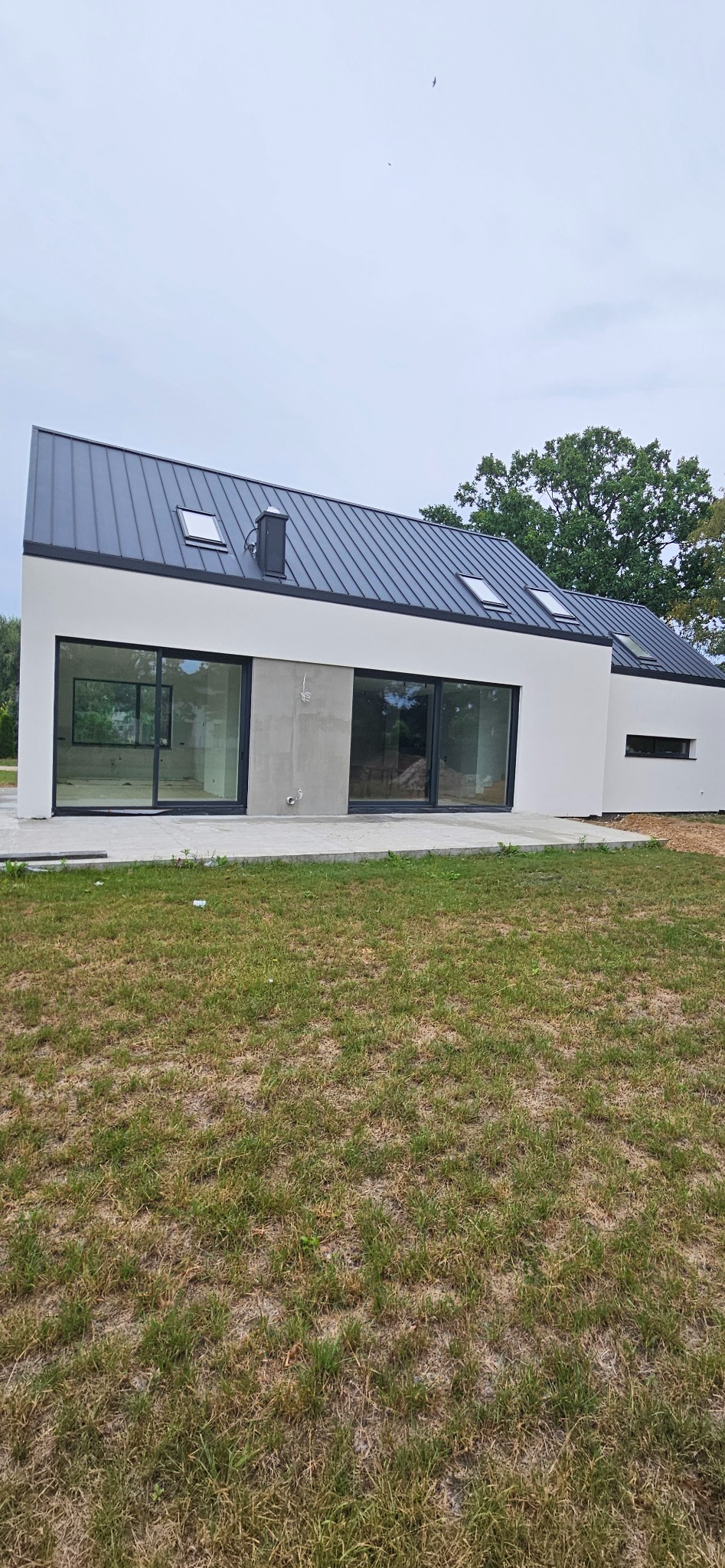 Nowoczesny, biały dom jednorodzinny z grafitowym dachem z blachy na tle zieleni, widoczne duże okna przesuwne i fragment elewacji z surowego betonu.