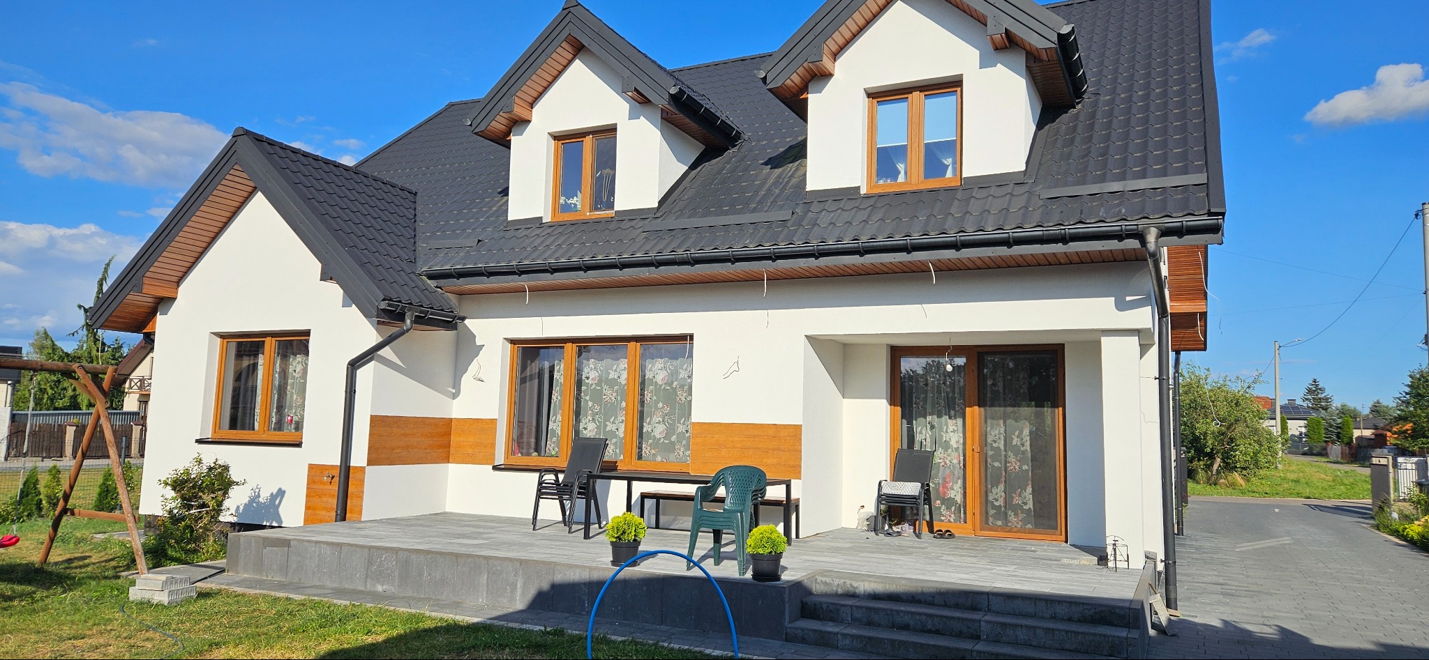 Biały dom jednorodzinny z czarnym dachem i drewnianymi akcentami elewacji, widoczne okna z drewnianą stolarką, taras z szarą kostką i meblami ogrodowymi, w tle błękitne niebo.