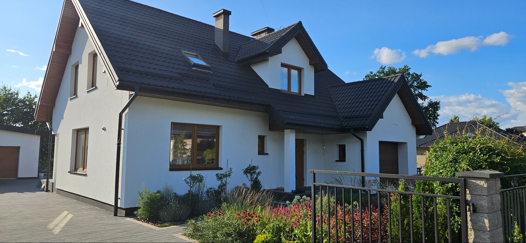 Nowoczesny dom jednorodzinny z białą elewacją, ciemną dachówką i zadbanym ogrodem z widocznym ogrodzeniem z metaloplastyki.