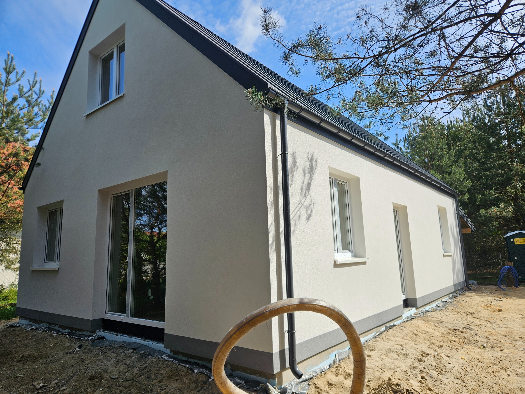 Świeżo otynkowany dom jednorodzinny z czarną dachówką, widok z zewnątrz, w tle drzewa. Wokół domu piasek i fragment węża ogrodowego.