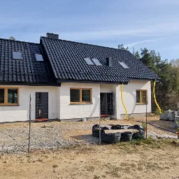 Nowo wybudowany dom z białą elewacją, czarnym dachem z dachówką i drewnianymi ramami okiennymi, z żółtymi rurami spustowymi i tymczasowym ogrodzeniem z siatki, na działce z piaskiem i gruzem,...