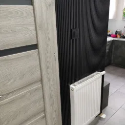 Drewnopodobne drzwi wewnętrzne w odcieniach szarości, czarna ściana z pionowych listew z włącznikiem światła oraz biały grzejnik panelowy, fragment kuchni w tle.