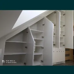 Biała zabudowa szaf z otwartymi drzwiczkami, dopasowana do skosu dachu, widoczne półki i zawiasy.