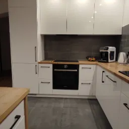 Nowoczesna kuchnia z białymi, lakierowanymi frontami szafek, drewnianym blatem i czarnymi, podłużnymi uchwytami, z widocznym piekarnikiem i ekspresem do kawy.