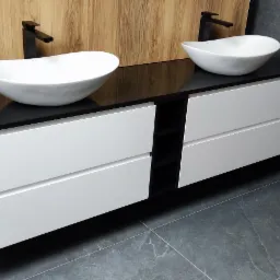 Nowoczesna umywalka nablatowa z czarną armaturą i białymi szafkami z szufladami, na tle drewnianej ściany i szarych płytek podłogowych w łazience.
