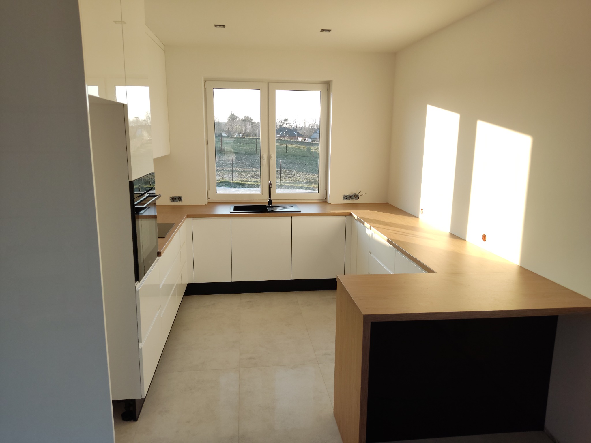 Nowoczesna, jasna kuchnia z białymi, lakierowanymi frontami i drewnianym blatem, widok na okno z krajobrazem, minimalistyczny styl.