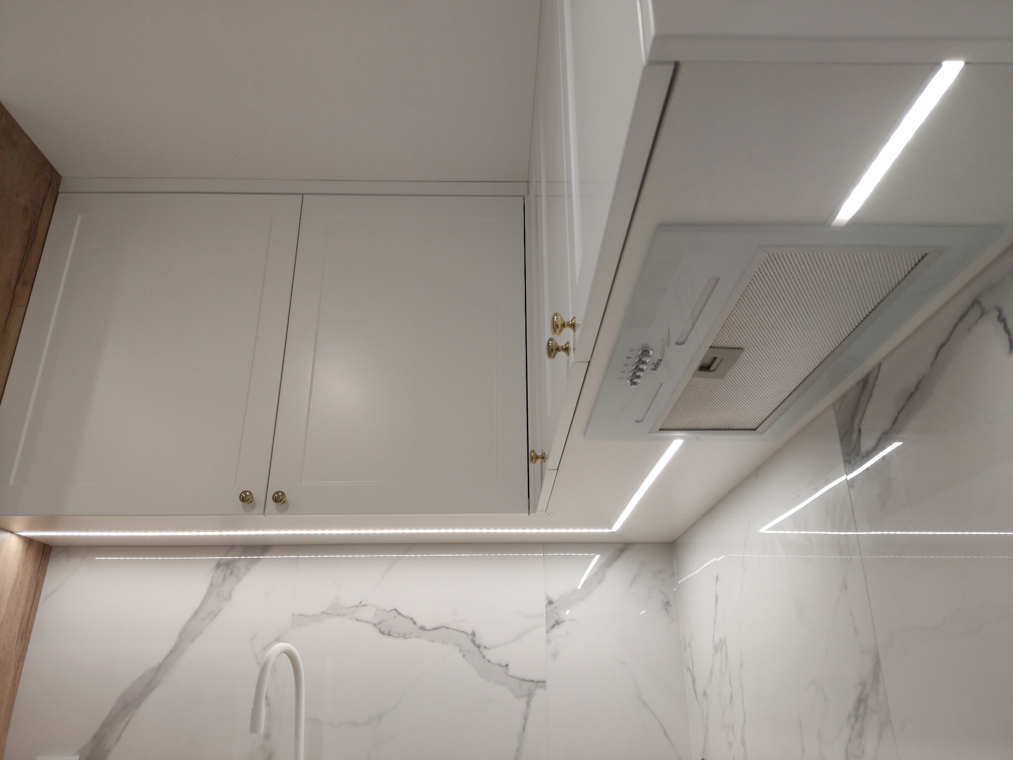 Białe szafki kuchenne z podświetleniem LED, złote gałki, marmurowy wzór na ścianie, nowoczesny okap kuchenny.