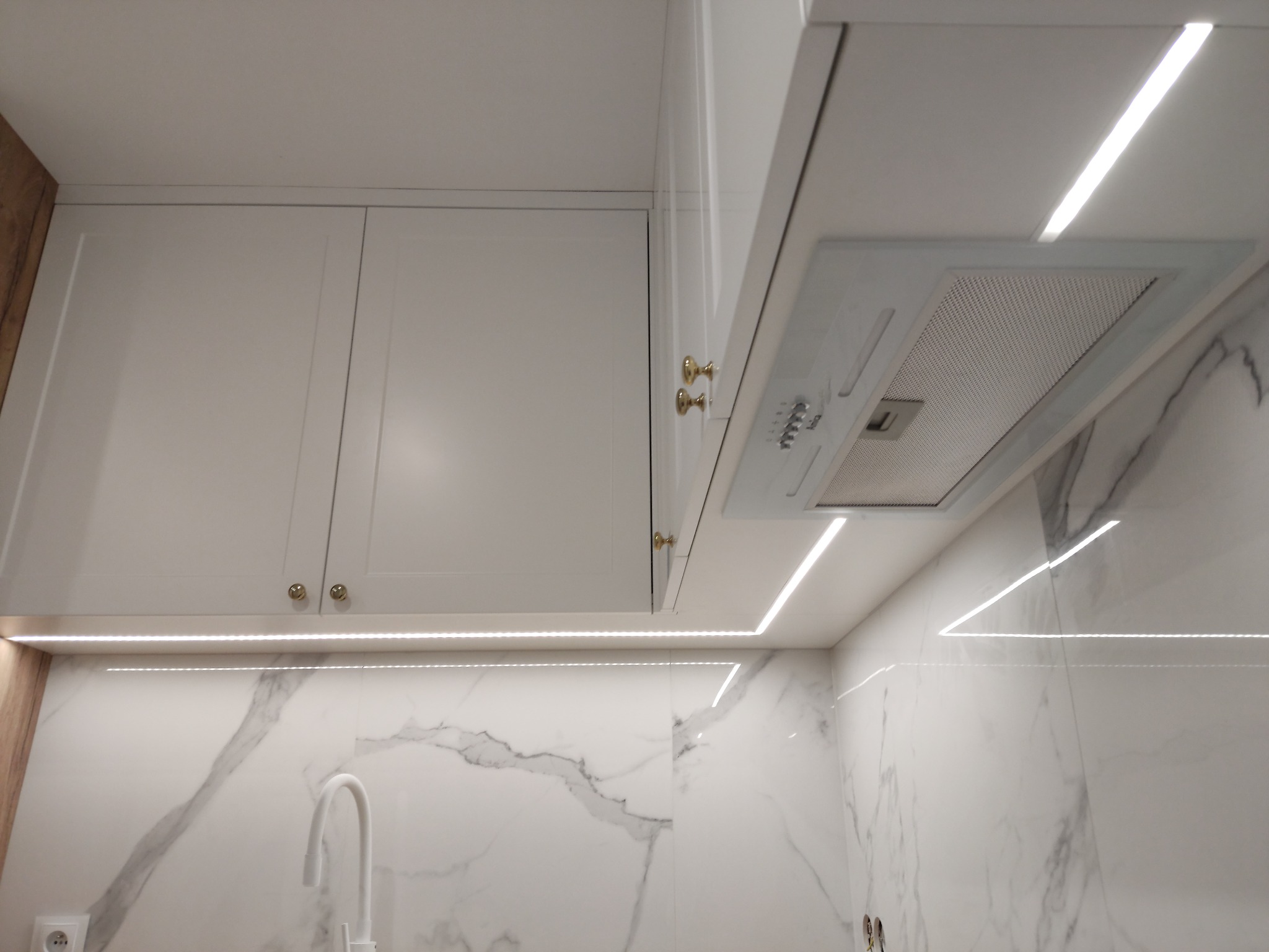 Białe szafki kuchenne z oświetleniem LED pod szafkami i marmurową ścianą, widoczny fragment okapu i bateria kuchenna.