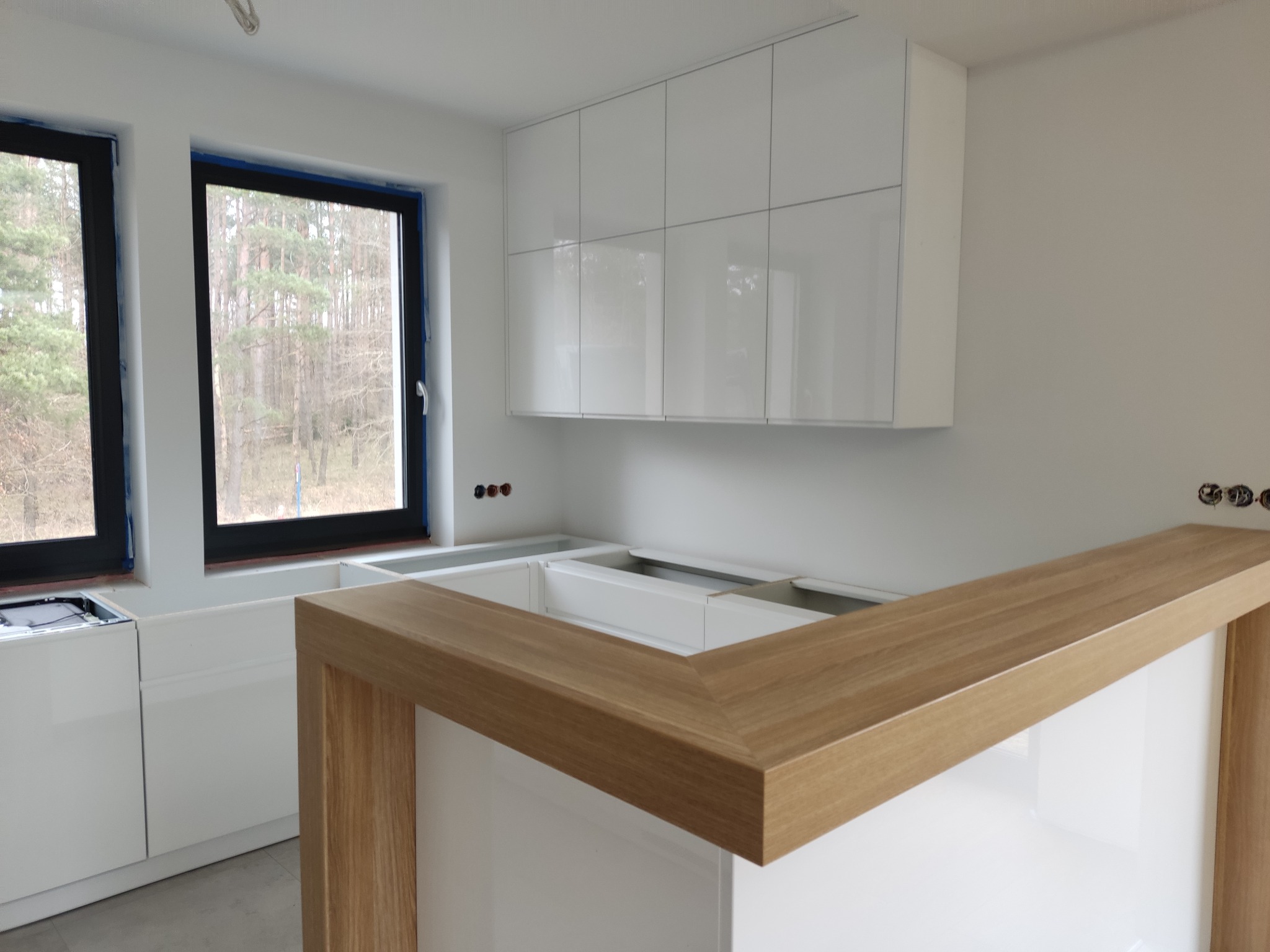 Wnętrze kuchni w trakcie montażu: białe szafki wiszące, drewniany blat baru z widocznymi niedokończonymi instalacjami elektrycznymi i oknem z widokiem na las.