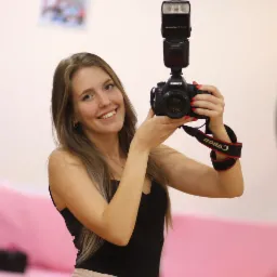 Uśmiechnięta blondynka robiąca selfie profesjonalnym aparatem Canon z lampą błyskową na tle jasnoróżowej ściany.