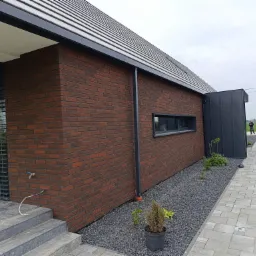 Ceglana elewacja domu jednorodzinnego z wąskim oknem, czarną rynną i szarą kostką brukową na podwórku.