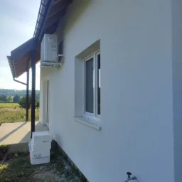 Świeżo otynkowana elewacja domu jednorodzinnego z widocznym klimatyzatorem, białymi oknami i stosem styropianu przy fundamencie. Widoczny kran na ścianie i fragment zadaszonego tarasu.