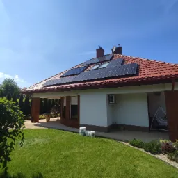 Dom jednorodzinny z panelami słonecznymi na czerwonym dachu, widoczny z zadbanego trawnika, z pojemnikami na farbę przy ścianie.