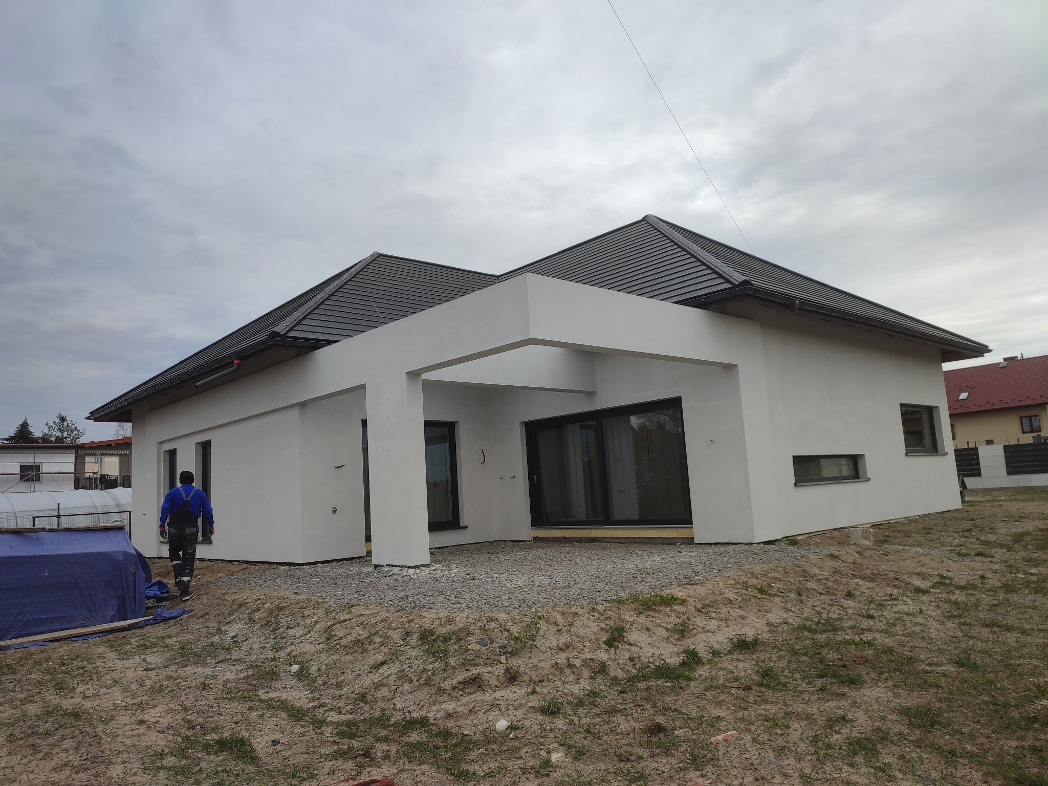 Nowoczesny dom jednorodzinny z płaskim dachem i białą elewacją, widok z zewnątrz, pracownik w oddali w niebieskim kombinezonie, teren wokół domu w trakcie prac wykończeniowych, pochmurne niebo.