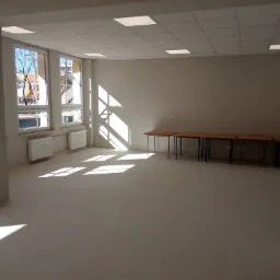 Jasne, puste pomieszczenie biurowe po remoncie, z widocznymi grzejnikami pod oknami, rzędami stołów i światłem słonecznym wpadającym przez okna.