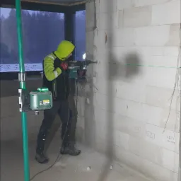 Elektryk w żółtej bluzie roboczej wierci otwór w ścianie z betonu komórkowego w surowym wnętrzu budynku, używając wiertarki udarowej i poziomicy laserowej.