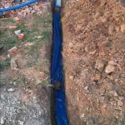 Wykop z niebieską folią ochronną wzdłuż fundamentu budynku w trakcie budowy, widoczne wystające rury instalacyjne.