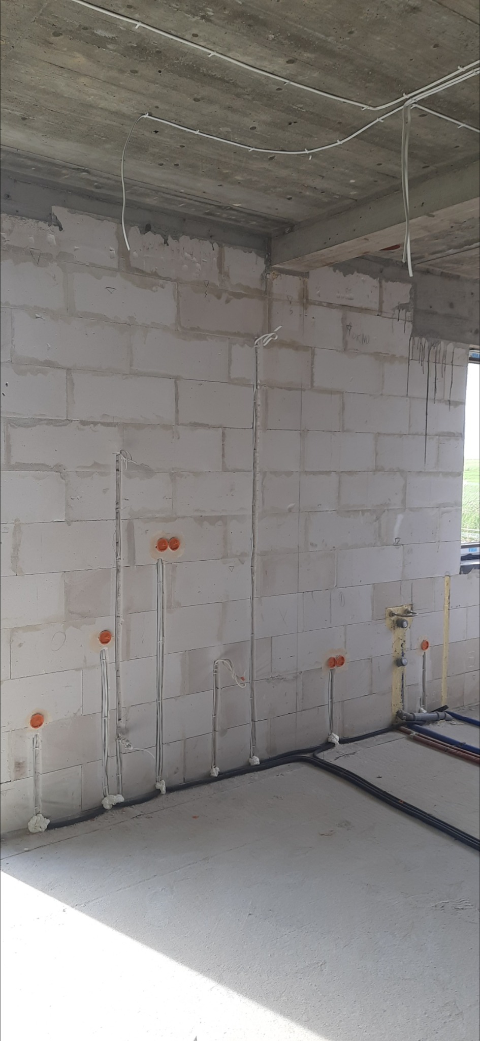 Surowy stan instalacji elektrycznej w budynku z białego bloczka: pionowe kable w pianie montażowej prowadzą do pomarańczowych puszek, poziome kable w czarnych rurkach przy podłodze, przewody...