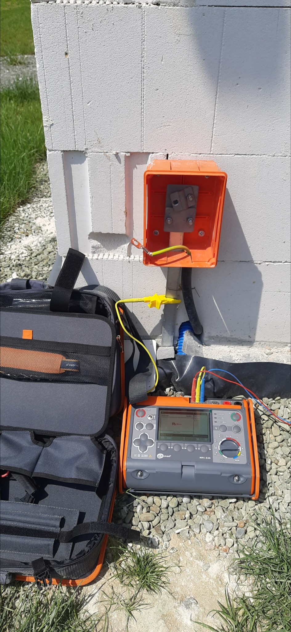Pomarańczowa puszka elektryczna z przyłączonym uziemieniem, obok torba narzędziowa i miernik elektryczny Sonel MPI-525 na tle ściany z betonu komórkowego i żwiru.