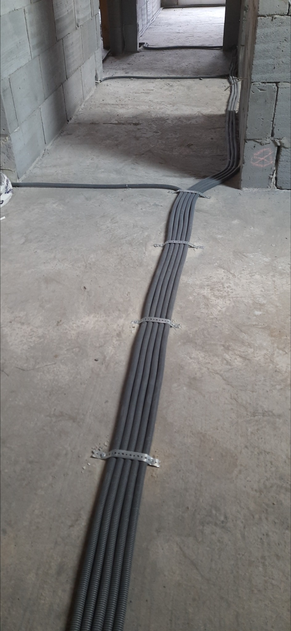 Sześć szarych rur karbowanych ułożonych równolegle, przymocowanych metalowymi obejmami do surowego betonowego podłoża w trakcie instalacji elektrycznej w budynku w stanie deweloperskim.