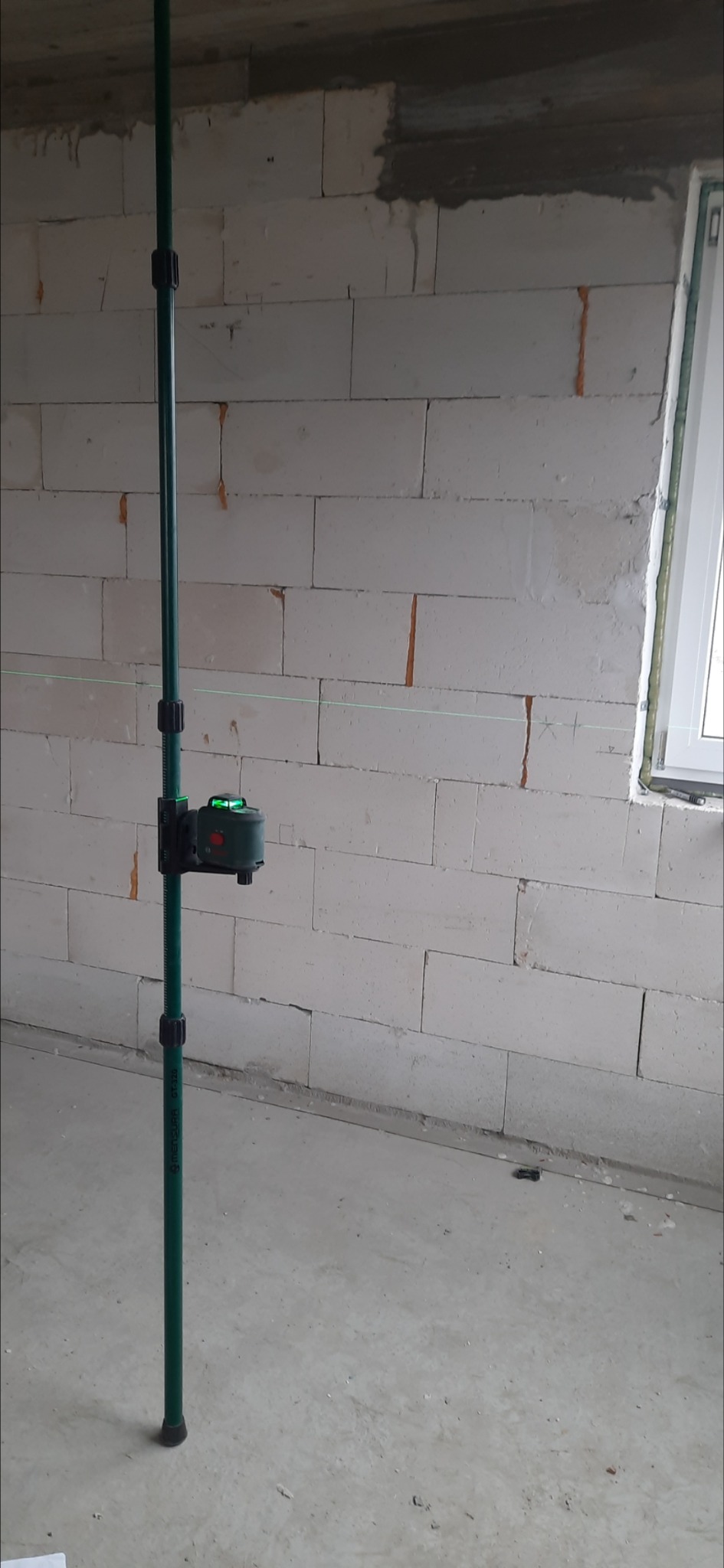 Zielony laser krzyżowy ustawiony na pionowej tyczce, wyświetlający poziomą wiązkę światła na ścianie z białych pustaków w surowym wnętrzu budynku.