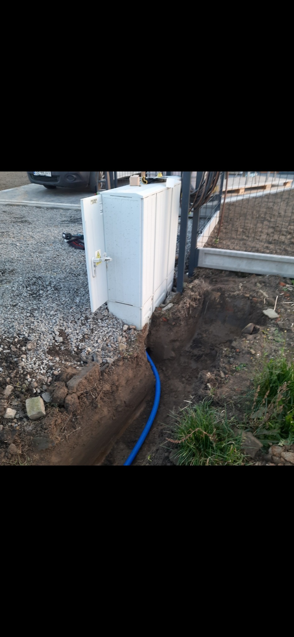 Otwarta szafka elektryczna przy ogrodzeniu, z wykopem i niebieską rurą kablową w ziemi, widoczny fragment samochodu w tle.