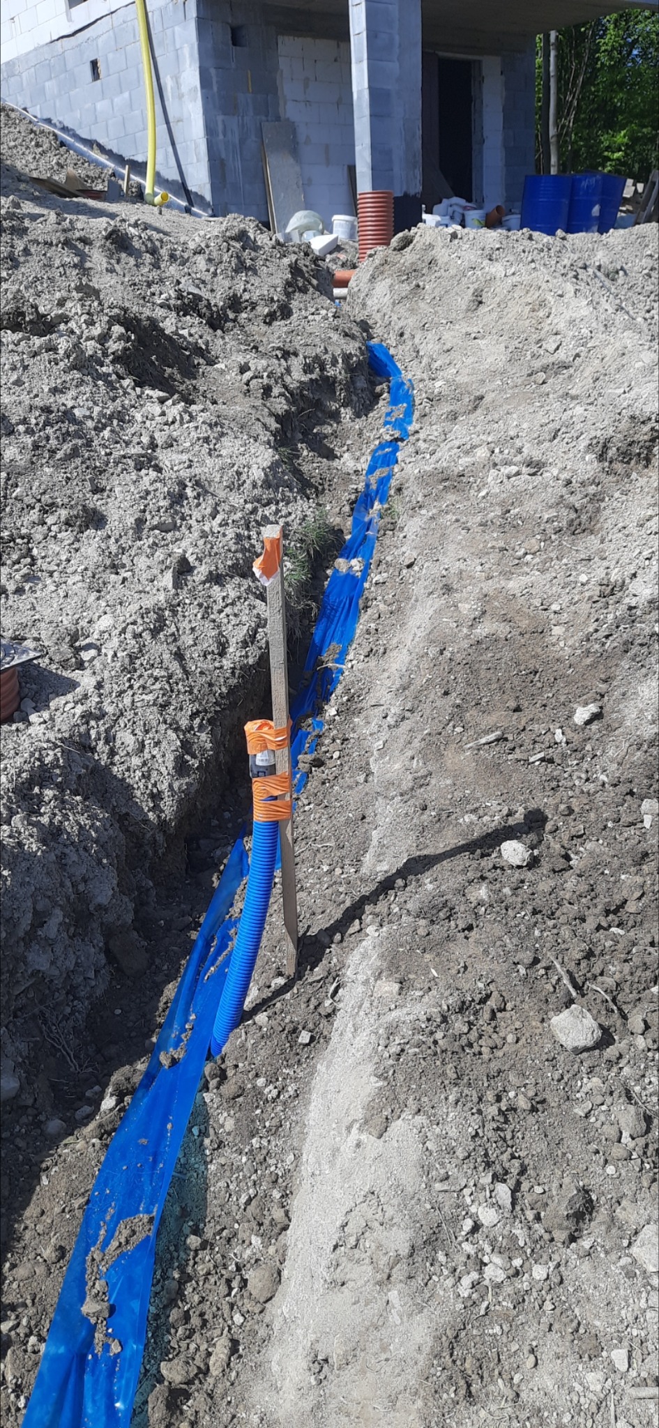 Niebieski karbowany przewód elektryczny ułożony w wykopie, zabezpieczony folią, umocowany do drewnianego palika taśmą ostrzegawczą, w tle budynek w stanie surowym otwartym.